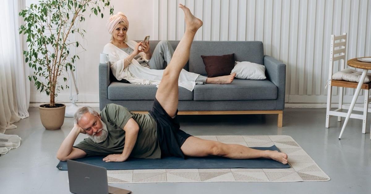 Older man doing a side-lying leg raise at home while following an exercise video on a laptop